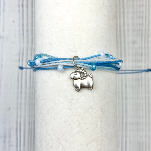 Op de afbeelding: Een armband met meerdere strengen in blauwe en witte tinten, versierd met kleine witte kralen. Een zilverkleurige ram-bedel hangt aan de armband. De armband is gewikkeld rond een wit cilindrisch object.