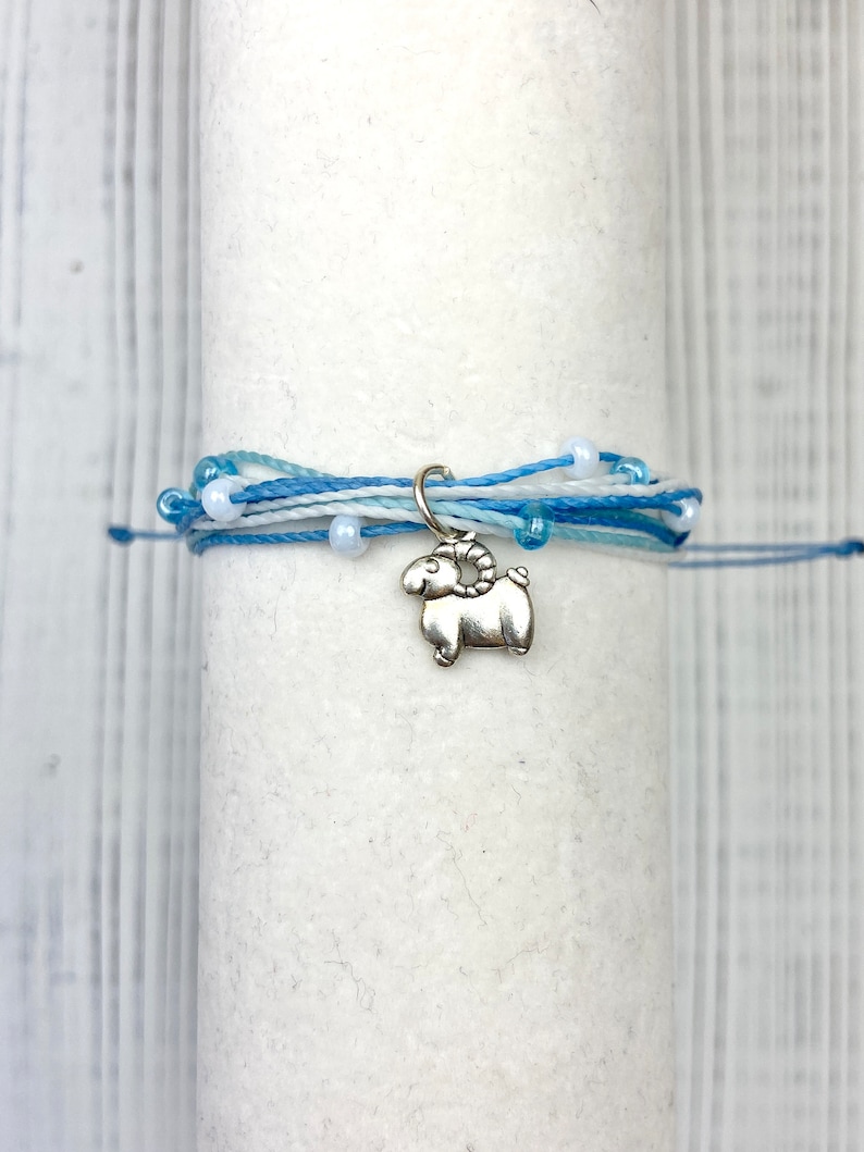 Op de afbeelding: Een armband met meerdere strengen in blauwe en witte tinten, versierd met kleine witte kralen. Een zilverkleurige ram-bedel hangt aan de armband. De armband is gewikkeld rond een wit cilindrisch object.