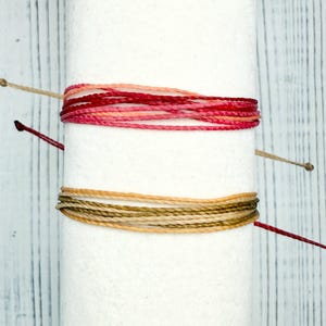 May include: Two sets of colorful bracelets are wrapped around a white cylinder. The top set features shades of red and pink, while the bottom set displays earth tones of tan and brown. The bracelets are made of multiple strands of thread.