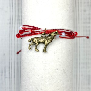 May include: Red and white string bracelet with a wolf charm. The charm is made of metal and has a detailed design.