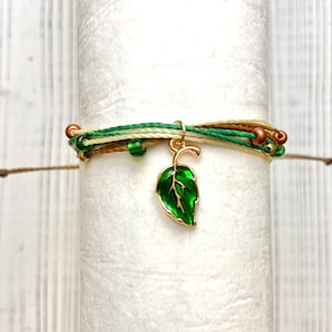 May include: A green leaf charm hangs from a multi-strand bracelet with brown, green, and white threads and green beads.