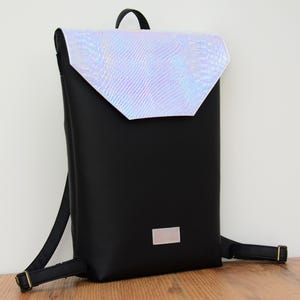 May include: A black backpack with a iridescent, snakeskin-textured flap. The flap has a white border and a white rectangular label on the front of the bag.