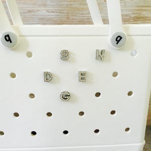 Peut inclure: Sac fourre-tout blanc avec une grille de trous et des strass qui &eacute;pellent le mot "SEND" au centre. Le sac a deux poign&eacute;es avec un bouton blanc sur chaque poign&eacute;e qui dit "Q".