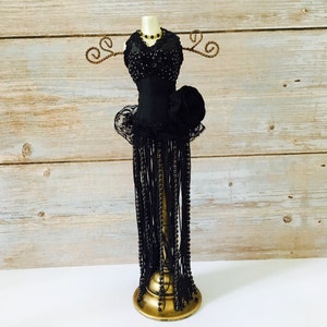May include: Black jewelry stand with a beaded skirt and a black bodice with a rose accent. The stand is made of metal and has a gold base.