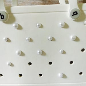 May include: White tote bag with a perforated design and white pearl embellishments. The bag has two handles with black letter 'a' and 'b' charms.