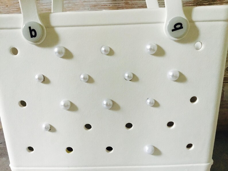 May include: White tote bag with a perforated design and white pearl embellishments. The bag has two handles with black letter 'a' and 'b' charms.