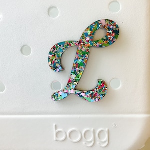 May include: A colorful, glittery letter L charm, perfect for adding a personalized touch to a white Crocs shoe. The charm is made of a durable material and features a variety of colors, including pink, blue, green, and yellow. The charm is attached to the shoe with a small hole.