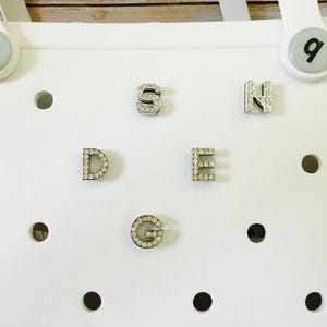 Peut inclure: Une surface blanche avec six breloques argent&eacute;es en forme de lettres S, D, E, N, G et un petit charme avec la lettre q. Les breloques sont d&eacute;cor&eacute;es de petits strass transparents.