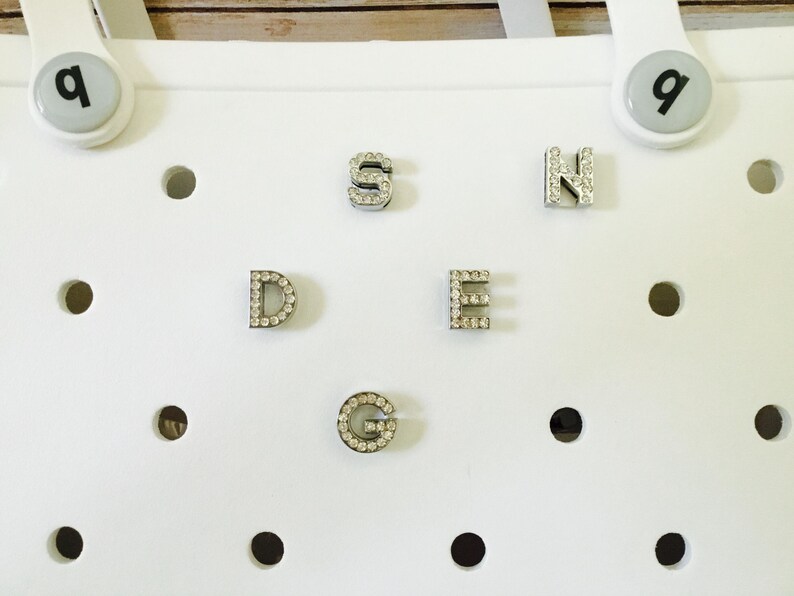 Peut inclure: Une surface blanche avec six breloques argent&eacute;es en forme de lettres S, D, E, N, G et un petit charme avec la lettre q. Les breloques sont d&eacute;cor&eacute;es de petits strass transparents.