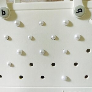 May include: White tote bag with a  'b' on each handle and a pattern of white pearl-like embellishments.
