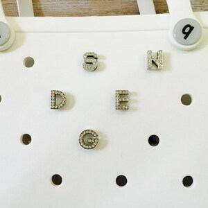 Peut inclure: Sac en plastique blanc avec une grille de trous. Les lettres D, E, G, N et S sont attach&eacute;es au sac. Chaque lettre est en m&eacute;tal et a de petits strass clairs autour des bords.