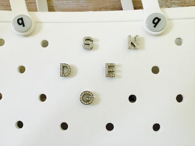 Peut inclure: Sac en plastique blanc avec une grille de trous. Les lettres D, E, G, N et S sont attach&eacute;es au sac. Chaque lettre est en m&eacute;tal et a de petits strass clairs autour des bords.