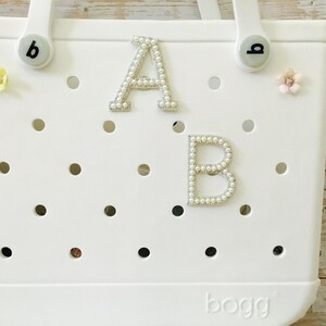 May include: White tote bag with the letters "A" and "B" embellished with faux pearls. The bag has a perforated design and is decorated with small floral accents. The brand name "bogg" is visible at the bottom.