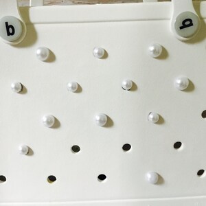 May include: White tote bag with a grid pattern and white pearl embellishments. The bag has two handles with black letter labels "a" and "b".