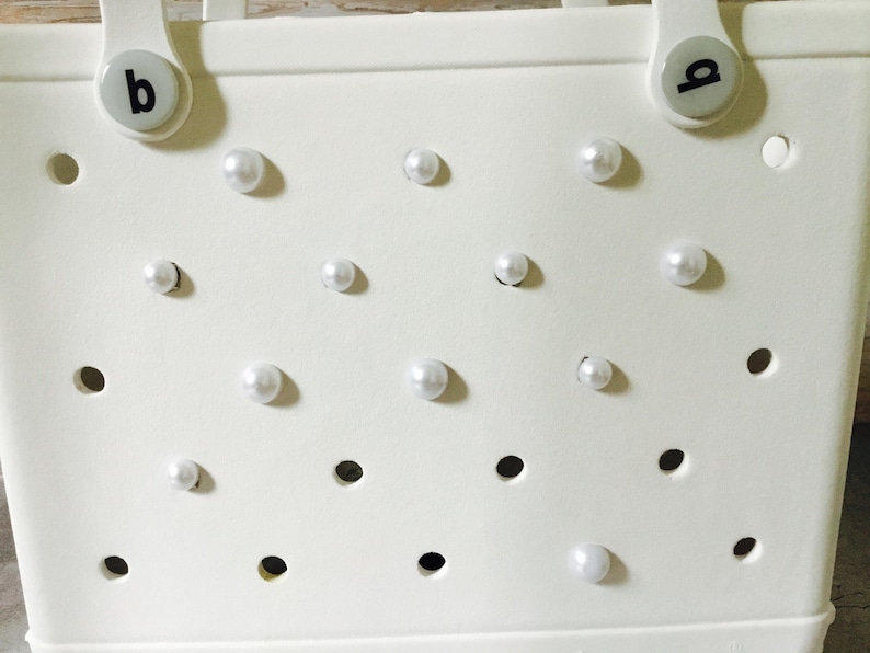 May include: White tote bag with a grid pattern and white pearl embellishments. The bag has two handles with black letter labels "a" and "b".