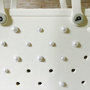 May include: White tote bag with a perforated design and white pearl embellishments. The bag has two handles and a 'b' logo on each side.