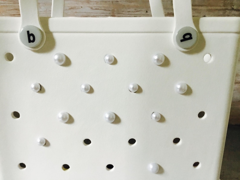 May include: White tote bag with a perforated design and white pearl embellishments. The bag has two handles and a 'b' logo on each side.