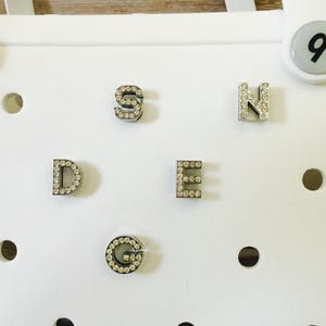 Peut inclure: Cinq breloques en forme de lettres argent&eacute;es avec des strass. Les lettres sont D, E, G, N et S.