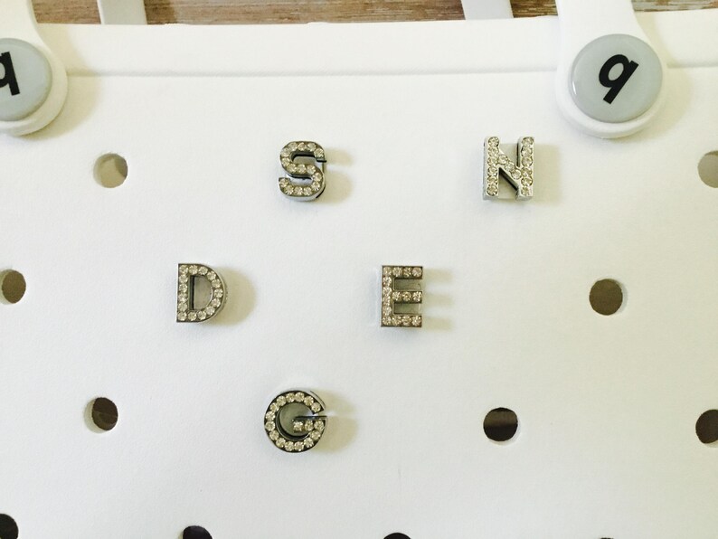 Peut inclure: Cinq breloques en forme de lettres argent&eacute;es avec des strass. Les lettres sont D, E, G, N et S.
