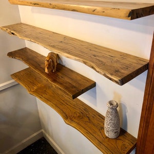May include: Four wooden floating shelves with live edges are mounted on a white wall. The shelves are different shades of brown and have a natural, organic look. A small wooden bear figurine and a decorative vase are placed on the shelves.