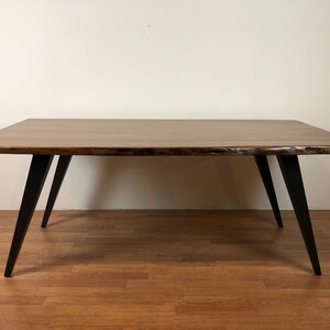 May include: A rectangular wooden table with a live edge top and black metal hairpin legs. The table is on a wood floor.