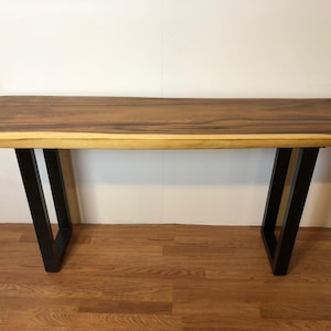 May include: A long, narrow wooden console table with a live edge top and black metal legs. The table has a natural wood finish and a rustic look.