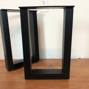 May include: Two black, rectangular metal table legs. The legs are made of thick, square tubing and have a modern, minimalist design. They are positioned on a wooden floor against a white wall.