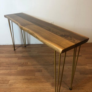 May include: A long, live edge walnut wood console table with a natural finish. The table has a unique, organic shape and is supported by four gold hairpin legs.