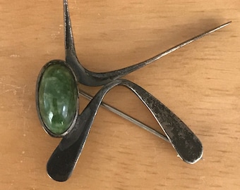 May include: A vintage silver-toned brooch with an abstract design. It features a large oval green stone set in a silver frame, with curved and pointed metal elements extending from it. The brooch has a pin closure on the back.