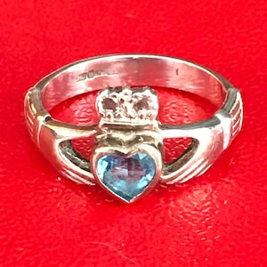 May include: A silver Claddagh ring with a blue heart-shaped gemstone. The ring features two hands clasped together, a heart, and a crown.