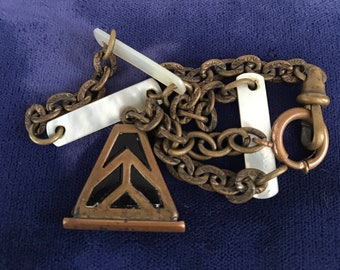 Op de afbeelding: Een messing kettingarmband met gegraveerde schakels, drie rechthoekige inlegstaafjes van wit parelmoer en een driehoekige hanger met zwart emaille chevron-detail. Het sieraad is voorzien van een ronde veerringsluiting en een draaibare haak.