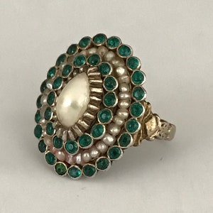 Op de afbeelding: Een zilveren ring met een grote witte parel omringd door een cirkel van groene edelstenen en kleinere parels.