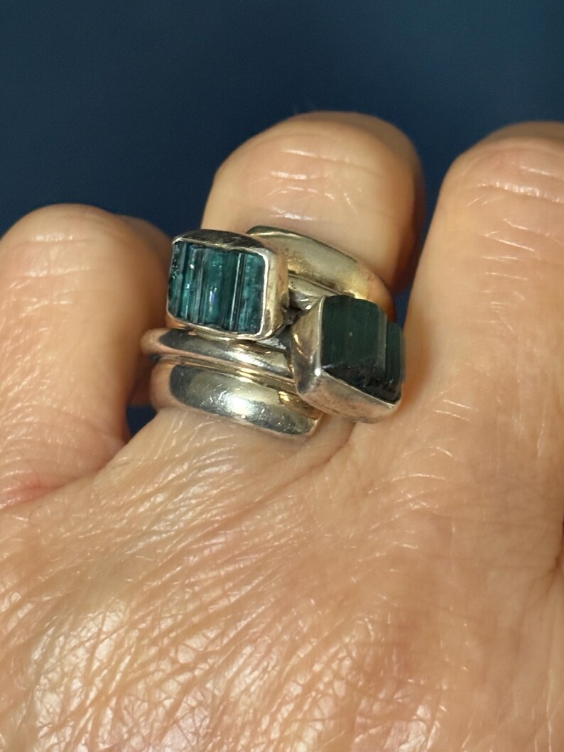 Peut inclure: Une bague en argent avec trois bandes et deux pierres pr&eacute;cieuses vertes. Les pierres pr&eacute;cieuses sont rectangulaires et pr&eacute;sentent un motif stri&eacute;.