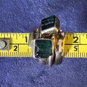 Peut inclure: Une bague en argent avec trois pierres vertes serties dans la bande. La bague est mesur&eacute;e &agrave; l'aide d'un m&egrave;tre ruban jaune.