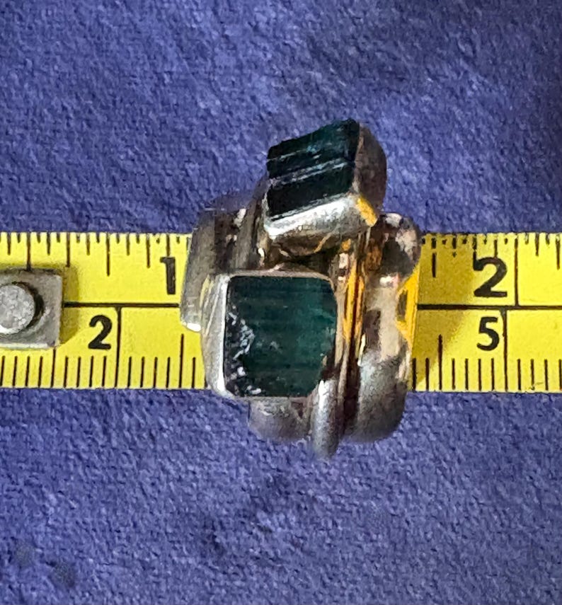 Peut inclure: Une bague en argent avec trois pierres vertes serties dans la bande. La bague est mesur&eacute;e &agrave; l'aide d'un m&egrave;tre ruban jaune.