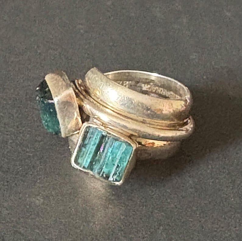 Peut inclure: Une bague en argent avec trois bandes, chacune sertie d'une pierre pr&eacute;cieuse verte diff&eacute;rente. La bande sup&eacute;rieure pr&eacute;sente une grande pierre pr&eacute;cieuse verte facet&eacute;e. La bande du milieu pr&eacute;sente une pierre pr&eacute;cieuse verte rectangulaire plus petite. La bande inf&eacute;rieure est unie.