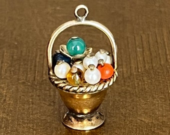 May include: A gold charm in the shape of a basket filled with colorful gemstones and pearls. The charm is perfect for adding a touch of whimsy to a necklace or bracelet.