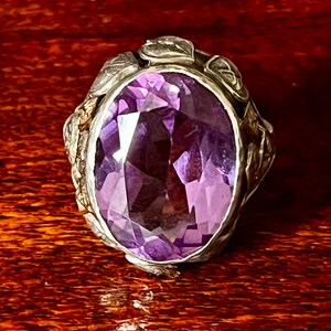 May include: A silver ring with an oval-shaped amethyst gemstone set in a floral design.