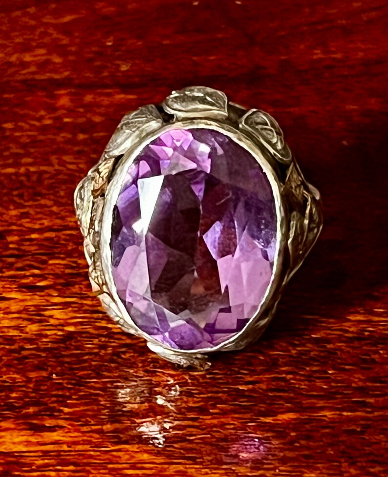 May include: A silver ring with an oval-shaped amethyst gemstone set in a floral design.