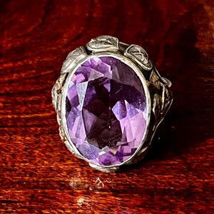 May include: A silver ring with an oval-shaped amethyst gemstone set in a floral design.