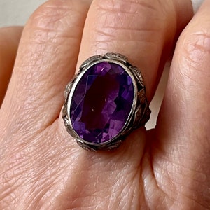 May include: A silver ring with an oval-shaped amethyst gemstone. The ring has a detailed floral design around the stone.