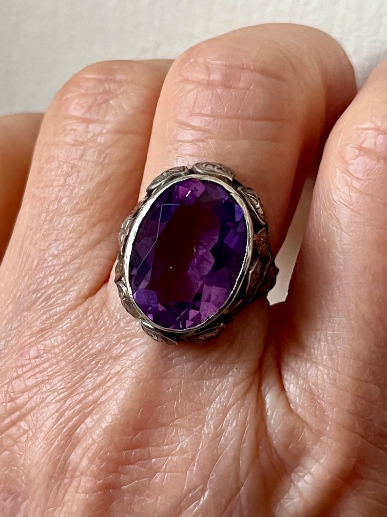 May include: A silver ring with an oval-shaped amethyst gemstone. The ring has a detailed floral design around the stone.