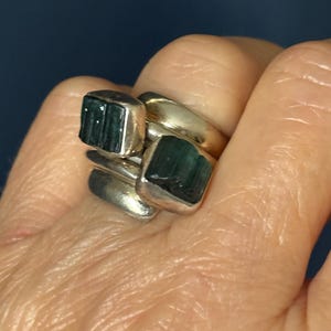 Peut inclure: Une bague en argent avec deux pierres pr&eacute;cieuses vertes serties dans la bande. La bague est port&eacute;e au doigt d'une personne.