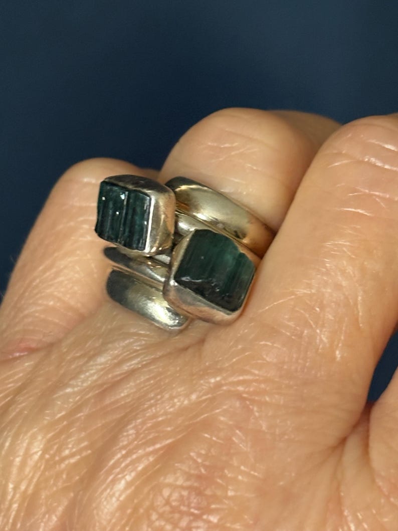 Peut inclure: Une bague en argent avec deux pierres pr&eacute;cieuses vertes serties dans la bande. La bague est port&eacute;e au doigt d'une personne.