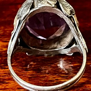 May include: A silver ring with an ornate design featuring leaves and a large, faceted amethyst gemstone.