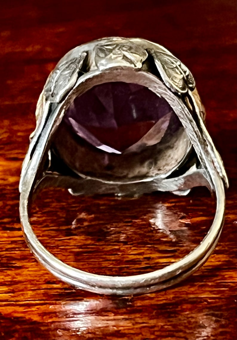May include: A silver ring with an ornate design featuring leaves and a large, faceted amethyst gemstone.