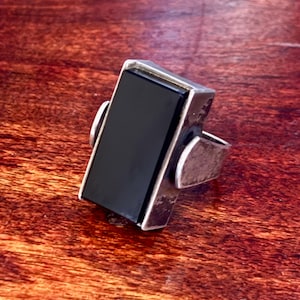Könnte beinhalten: Ein rechteckiger Silberring mit einem großen, schwarzen Onyxstein. Der Ring hat ein schlichtes, modernes Design mit poliertem Finish. Der Onyxstein ist in einer Zargenfassung gefasst, auf einer Holzoberfläche.