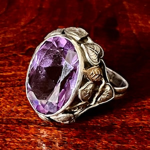May include: A silver ring with an oval-shaped amethyst gemstone set in a floral design.