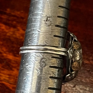 May include: A silver ring with a large, ornate design featuring gold accents. The ring is being measured on a metal ruler.