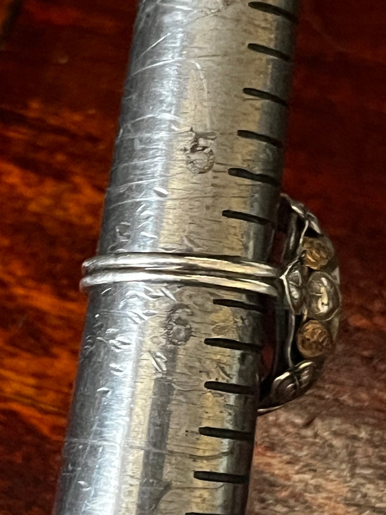 May include: A silver ring with a large, ornate design featuring gold accents. The ring is being measured on a metal ruler.